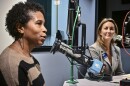 Two women sit inside a radio studio. They are seated at microphones. One on left (Brandy Williams) is talking while the other (Julie Donelon) is listening.