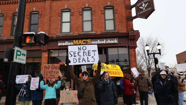 A small group of people stand in front of a crosswalk in downtown Saline. One woman in the center of the image holds a sign that reads “No Secret Deals.”