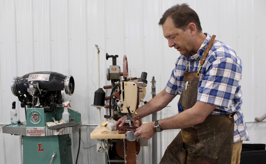 A man in a plaid shirt and apron uses an industrial sewing machine to stitch through leather.