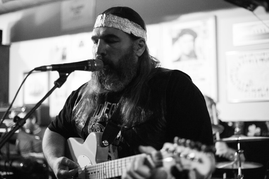 A black-and-white photo of a bearded man seen from the chest up playing guitar and singing into a microphone. He's wearing a headband made from tin foil.