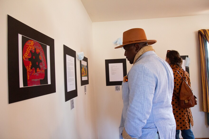 A photo of a man with brown skin wearing a brimmed hat, a light blue suit and a scarf. He's looking at a framed type-written page on the wall. 