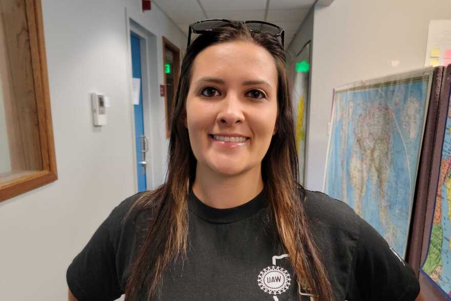 A woman stands in a hallway, smiling at the camera. She has brown hair, going down past her shoulders and sunglasses on her head. Her shirt has the UAW logo on it.