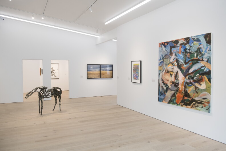 An art exhibit with white walls and pale wooden floors. There is a colorful painting on the wall closest to the camera and steel horse standing in the middle of the room.