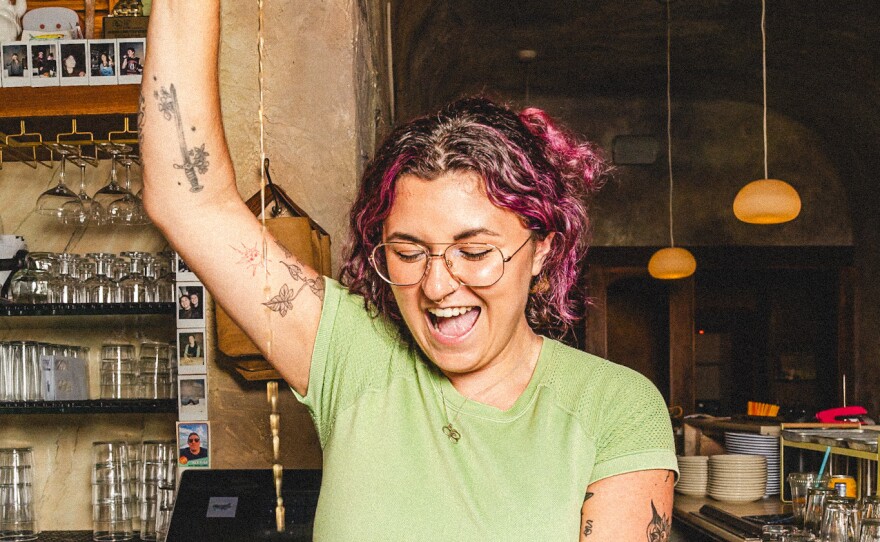 A smiling woman is bartending. She stands behind the bar pouring a drink from a cocktail shaker. She is wearing a cropped light green t-shirt, jeans and glasses