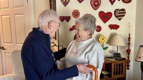 Elderly man stands with his cane and embraces his elderly wife, who smiles at him. They stand in a living room. A wall lined with pink and red wooden hearts is behind them.
