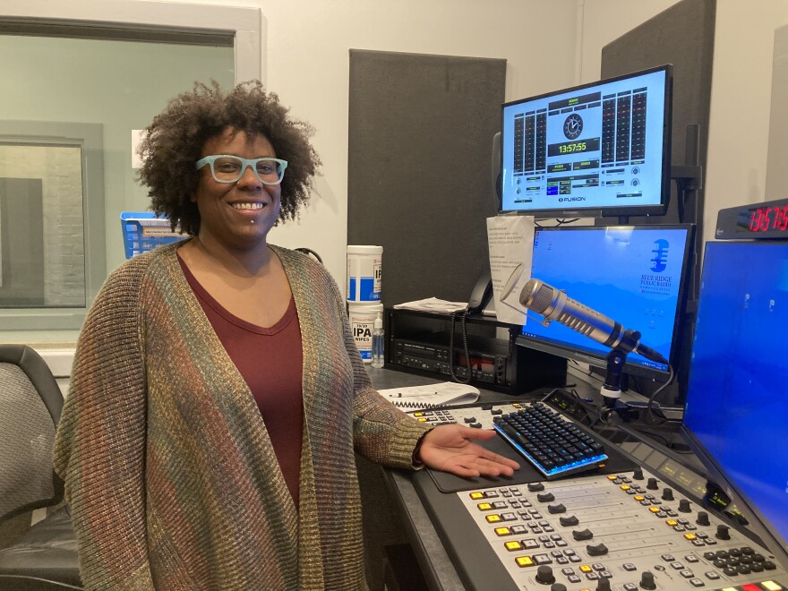 BPR Host Anastasia Marie, wearing a muted green, yellow and burgundy stripped cardigan and aqua glasses, stands before the on-air console, microphone and computers in one of BPR's studios.