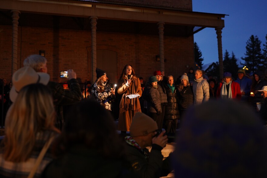 The Vice Mayor of Truckee dressed in a brown coat speaks into a microphone to a group of people outdoors in the evening at a vigil.