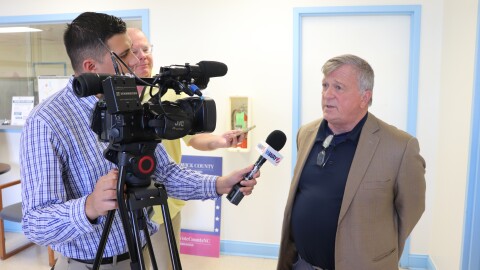 Rich Alt, an older white man with white hair in a tan sport coat, talks to a group of reporters inside the Board of Elections building. One reporter is filming him while holding a mic up to his face, the other is using his phone to record a video. 