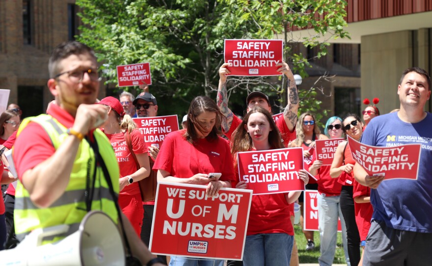 A group of people participate in an outdoor rally in support of University of Michigan nurses. In the foreground, a man in a neon safety vest speaks into a megaphone. Behind him, several individuals in red shirts hold up red and white signs that read "SAFETY STAFFING SOLIDARITY" and "WE SUPPORT U OF M NURSES." To the right, a man in a blue Michigan Medicine t-shirt holds a similar sign. The group is standing in a paved area with green trees and university buildings in the background.