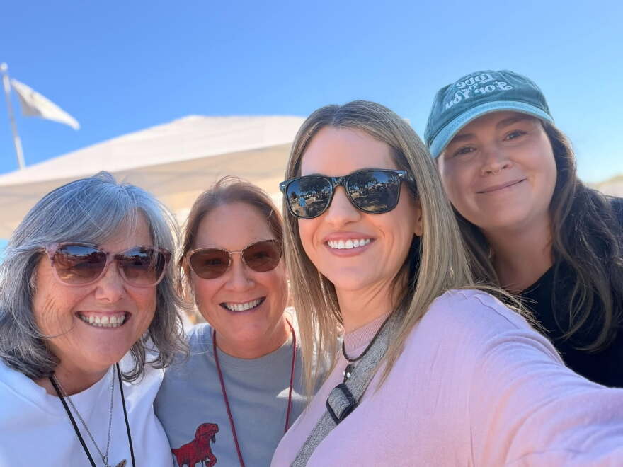 Four women smiling for a outdoor selfie under a bright blue sky with white event tents visible in the background; three wear sunglasses and one wears a baseball cap.