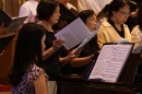 A woman plays a grand piano, behind which a choir sings