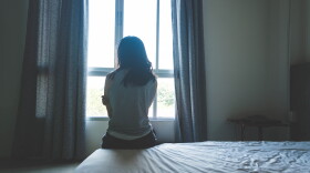 A woman's back is facing the viewer. She sits on a bed with crumpled sheets. The room has bare walls, curtains, and a night stand. The woman faces window. Light flows through the glass from the outside.