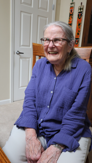 And elderly woman with glasses sits in a dining room chair, smiling. The room is sunny. Her gray hair is pulled back in a ponytail. She wears white pants, and a light purple blouse.