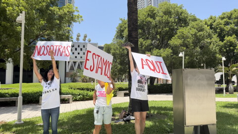 Anisley Cortes, Judith Castellano and Anett Uset-Dumont wave signs that read "NOSOTRAS ESTAMOS AQUI" (We are here) towards the windows of FDC Miami on July 15, 2026.