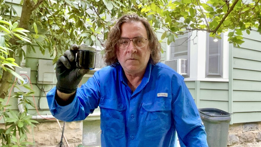 A man wearing a blue shirt and black gloves holds up a soil sample. He's standing in front a green house and a tree with green leaves.