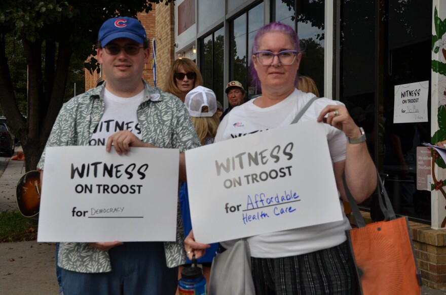 Two people stand on a sidewalk and hold signs that say witness on Troost.
