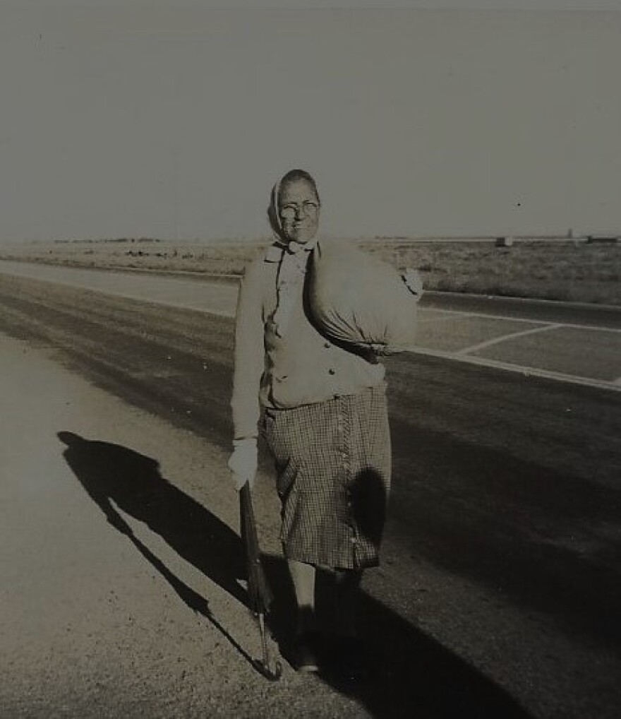 An old woman stands along a road with a pack slung over one shoulder. Her shadow stretches out behind her.