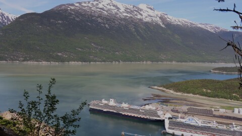 Two cruise ships docked in the foreground of a copper blue body of water, with snow-capped mountains on the opposite shore.