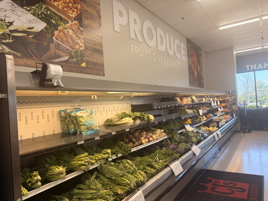 Produce is laid out in refrigerated shelves. A sign reading "Produce" is on the wall above.