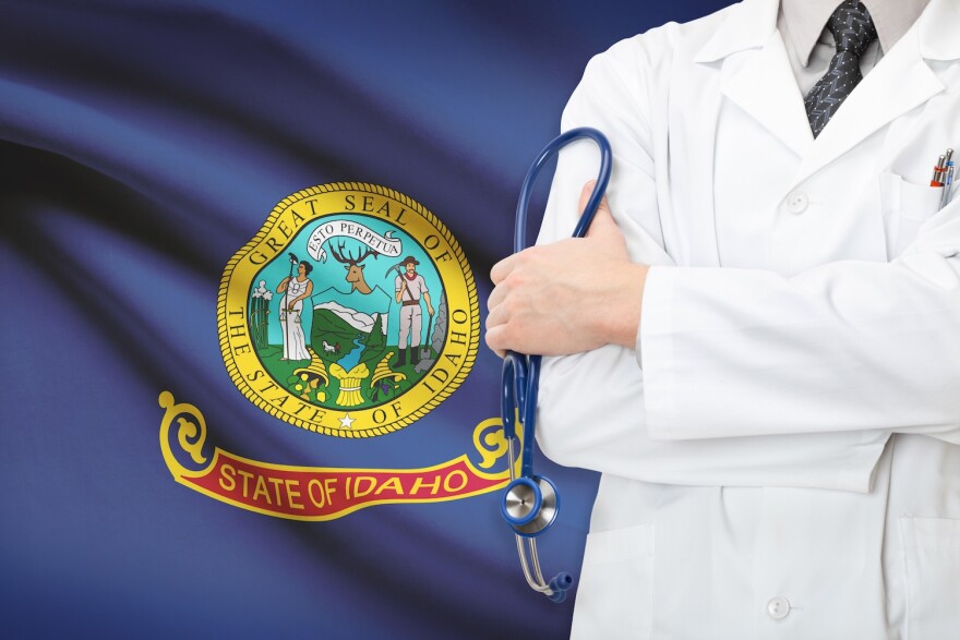 A person in a medical coat holding a stethescope, seen from shoulders to hips. The background is of the Idaho state flag.