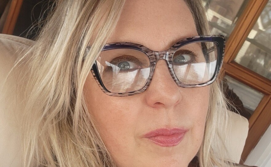 A smiling woman with blonde hair wearing thick glasses, a gold chain, and a rust-colored sweater with a white collar