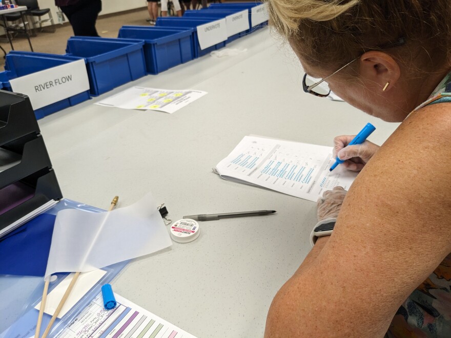 A women sitting at a table wearing disposable gloves is using a blue highlighter to make tally marks on a sheet of paper. There are blue bins on the table opposite of her.