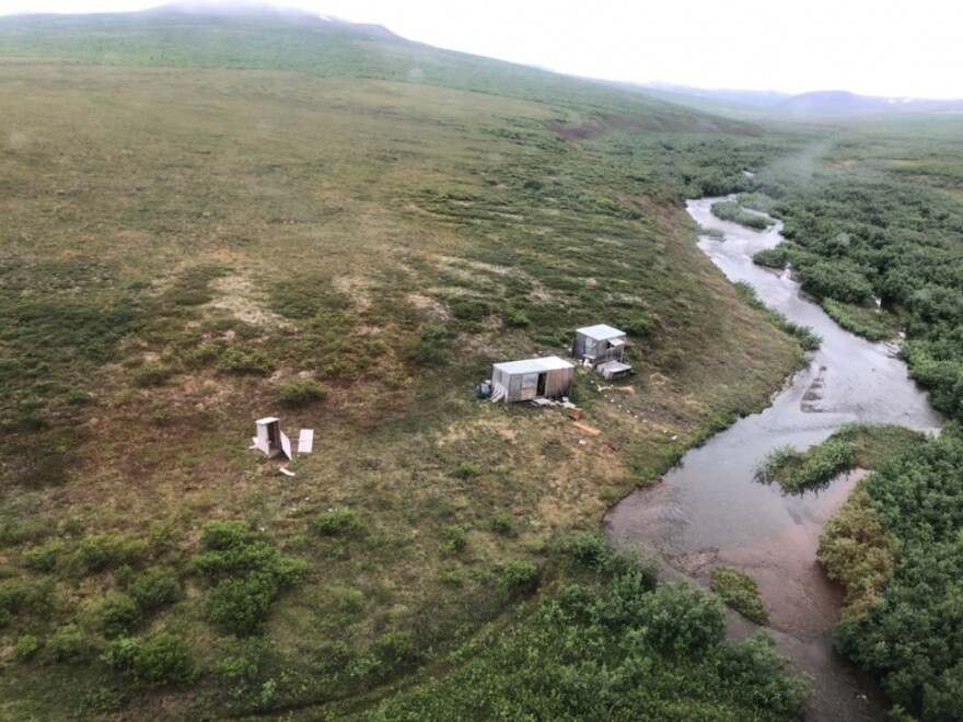 A photograph of a remote, grassy landscape with a few small shacks on it, near a river.