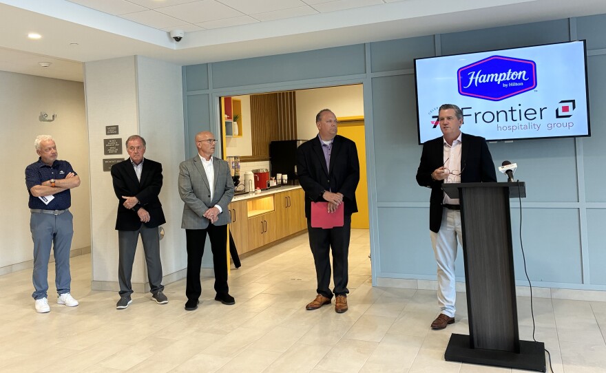 Five men stand in a hotel lobby, with one man speaking at a podium. Behind them is a wall-mounted screen displaying the Hampton by Hilton and Frontier Hospitality Group logos.