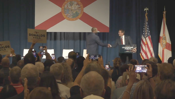 Florida Gov. Ron DeSantis shakes hands with U.S. Rep. Byron Donalds onstage in front of a large Florida flag as audience members watch and take photos.