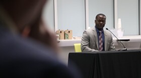Gilliam wears a suit and tie. He sits alone at a square table with a microphone. Gilliam looks toward the camera.