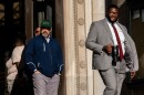 A Black man in a gray suit and red tie holds a black mug while walking out of a court building.
