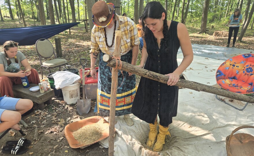 Cortney Collia instructs a rice camp attendee on how to dance in a jigging pit to help get the rice grain out of its husk. Large branches are tied together to provide support.