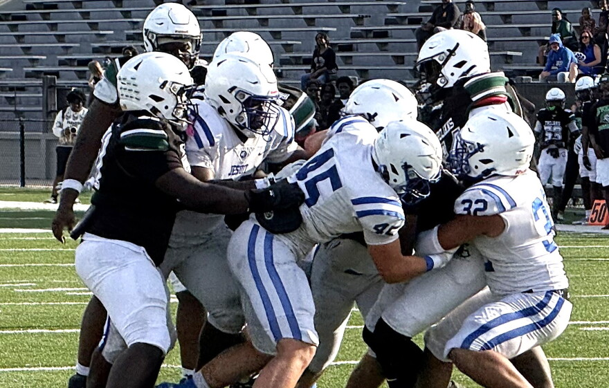 football action showing several players in a white uniform with blue strips tackling a player in a black jersey. both teams wearing white helmets. set on a green field with white football lines