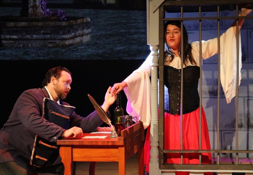 A woman in a white blouse, black corset and bright red skirt stands behind jail bars. She extends her right hand to a man seated at a desk. He is singing to her and wearing a military-looking uniform.