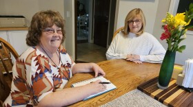 Two women sit at a table looking at the camera. The woman on the left has a pen in her right hand which is resting on a notebook. The second woman sits with her hands crossed on the table. There is a vase with yellow and red flowers on the table. 