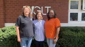 Three women posing for a photo in front of waist-high bushes and a red brick building with the letters WGLT on the front