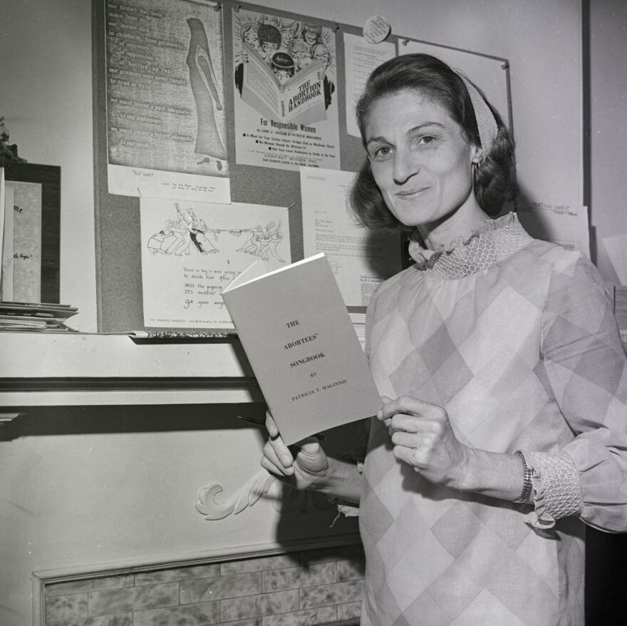 Patricia Maginnis holds her <em>Abortees' Songbook</em> and stands next to a bulletin board full of abortion information. The early abortion rights advocate, pictured here in 1970, died earlier this year.