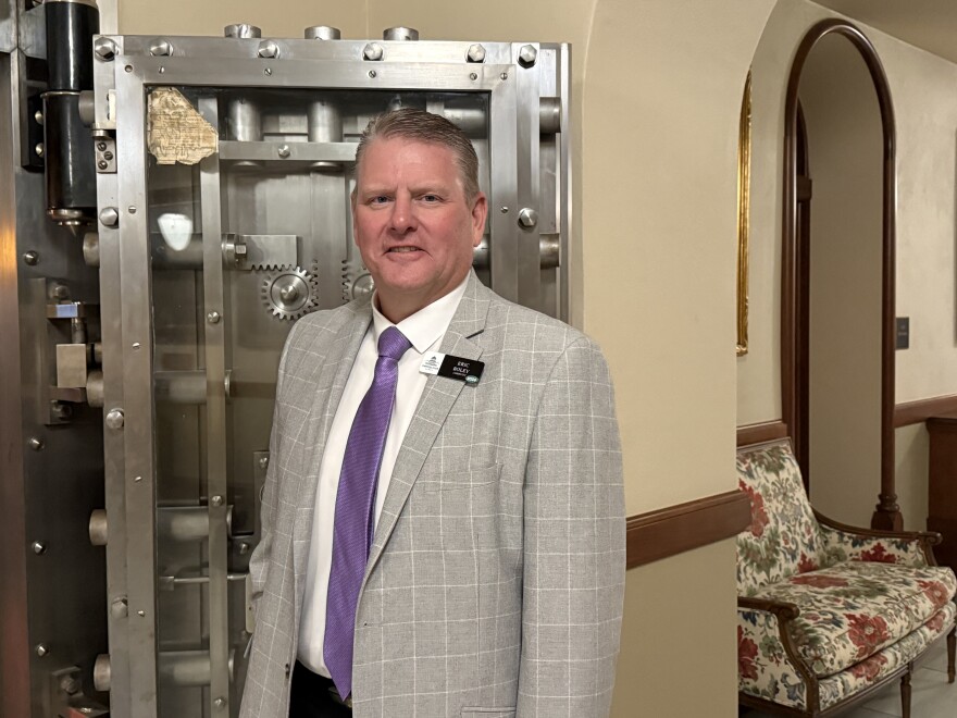 A man in a grey suit and purple tie stands in front of a metal door. 