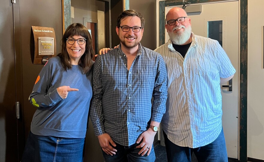 A woman and two men smile at the camera while standing in front of a recording studio.