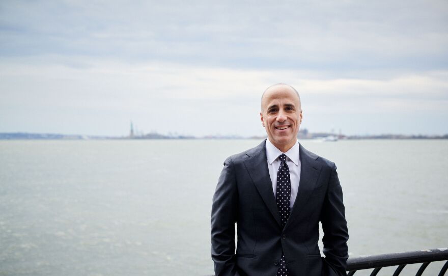 A smiling man with short grey hair wearing a black suit, white collared shirt, and black tie with white polka dots stands in front of a river with the Statue of Liberty in the background