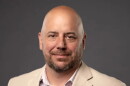 A bald man with a brown beard is wearing a beige blazer over a white plaid button-down shirt.