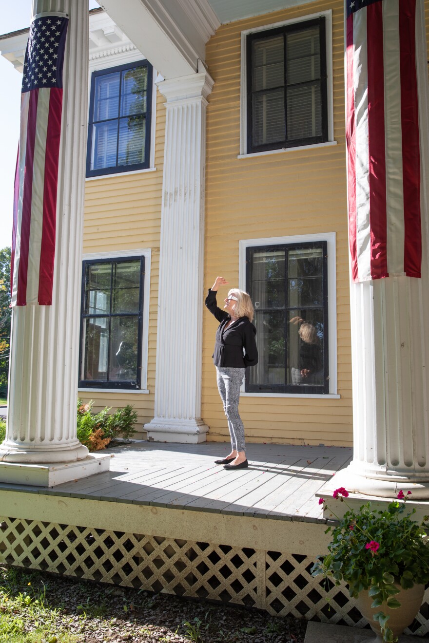 A woman stands on the front porch of what looks like a colonial home. She is facing left toward the sunlight, and she is shielding her face from the light while also wearing specialized glasses for viewing solar eclipses.