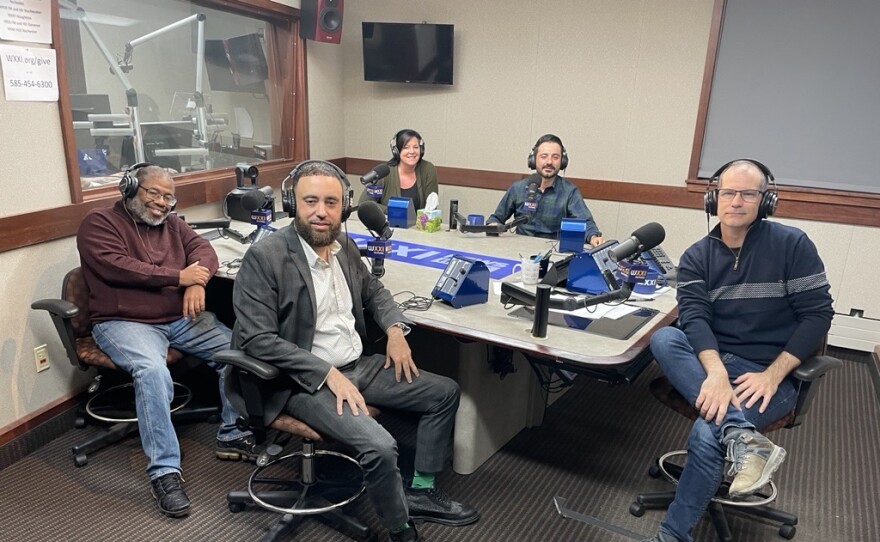 Five smiling people wearing headphones sit around a table in a radio talk studio: a man front left has short gray hair and a gray mustache and beard and is wearing glasses, a maroon sweater, jeans, and black shoes; a man front center has short dark hair and a dark mustache and beard and is wearing a white button-down shirt under a black jacket, black pants, and black shoes; a woman back left has short brown hair and is wearing a black top under a green sweater; a man back center has short dark hair and a dark mustache and beard and is wearing a blue button-down shirt; a man at right has very short dark hair and is wearing a blue pullover with gray horizontal stipes, jeans, and hiking shoes