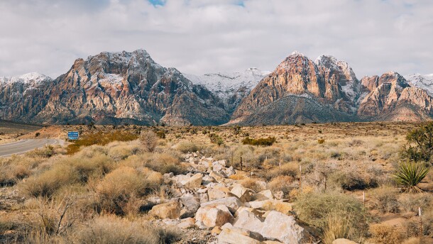 A desert landscape of dry scrub brush and scattered boulders stretches toward dramatic red and orange sandstone mountains dusted with snow at their peaks, under a cloudy winter sky, with a road visible on the left.