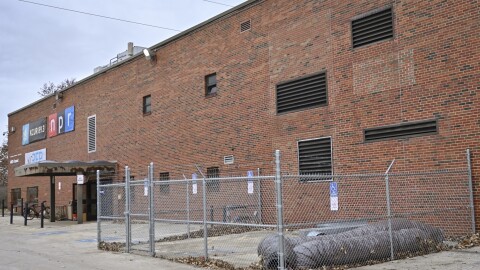 Exterior photo of a red brick, two story building with banners on the far left that read "KCUR 89.3," "NPR" and "KCREP." In the foreground  is a cyclone enclosure with some abandoned ductwork.