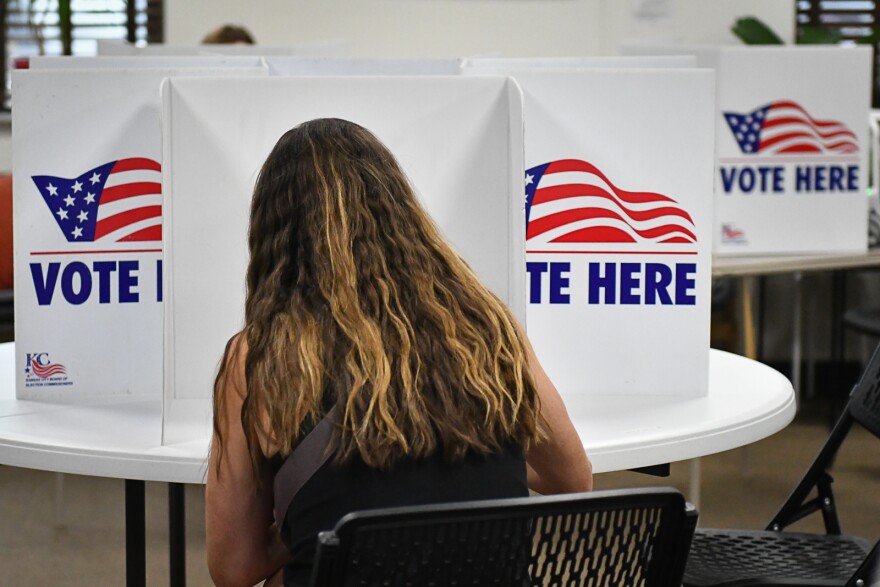 Interior photograph of a person sitting inside a room. They are seated in a folding chair at round table where there are "Vote Here" signs. They appear to be writing. 
