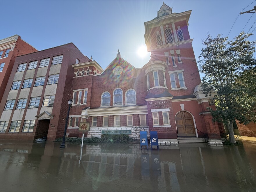 A red-brick church and adjacent buildings are surrounded by green-brown floodwater, about shin-deep.