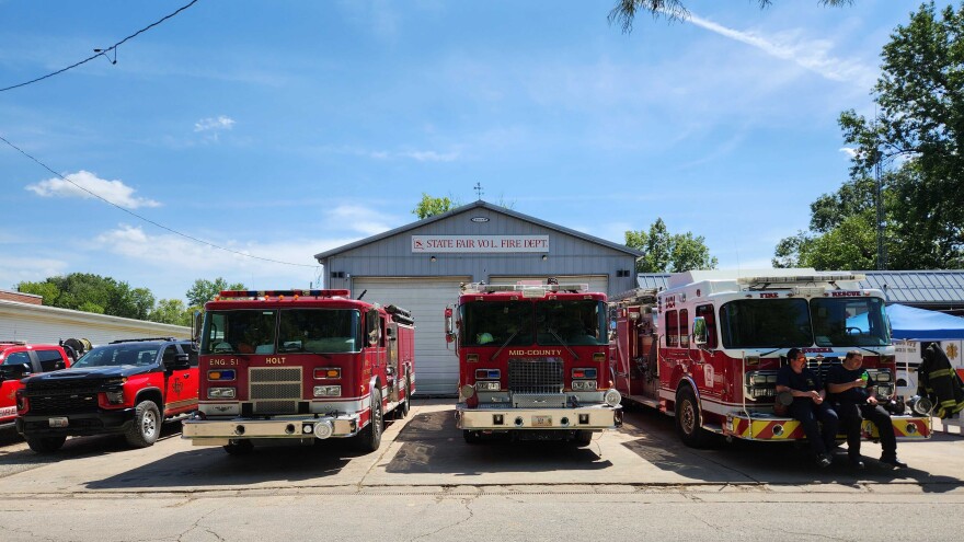 A building has a large sign on it that reads "State Fair Vol. Fire Dept." Several bright red fire engines and several pickup trucks sit in front of the building.
