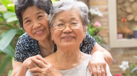 An elderly woman smiles at the camera with her daughter standing behind her.