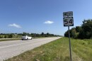 A white sedan drives down Highway 54. The photo is a wide shot of the road. A "West 54" road sign stands in the ditch. 